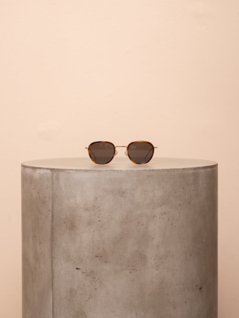 Brown wireframe sunglasses on pedestal against pink background.