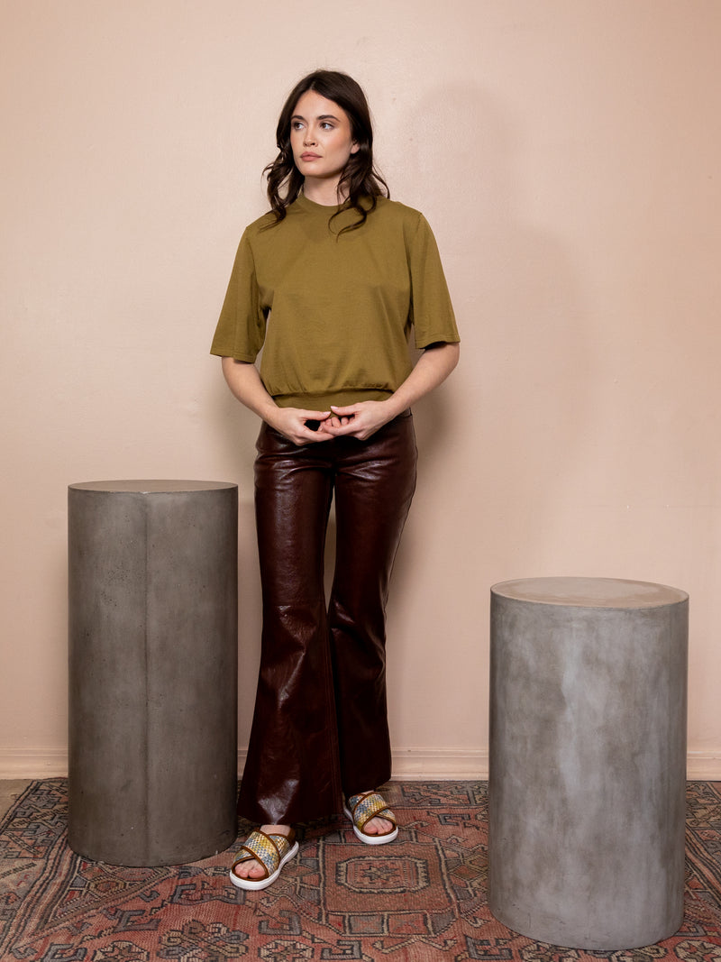 Woman wearing green top and leather brown pants against pink background.