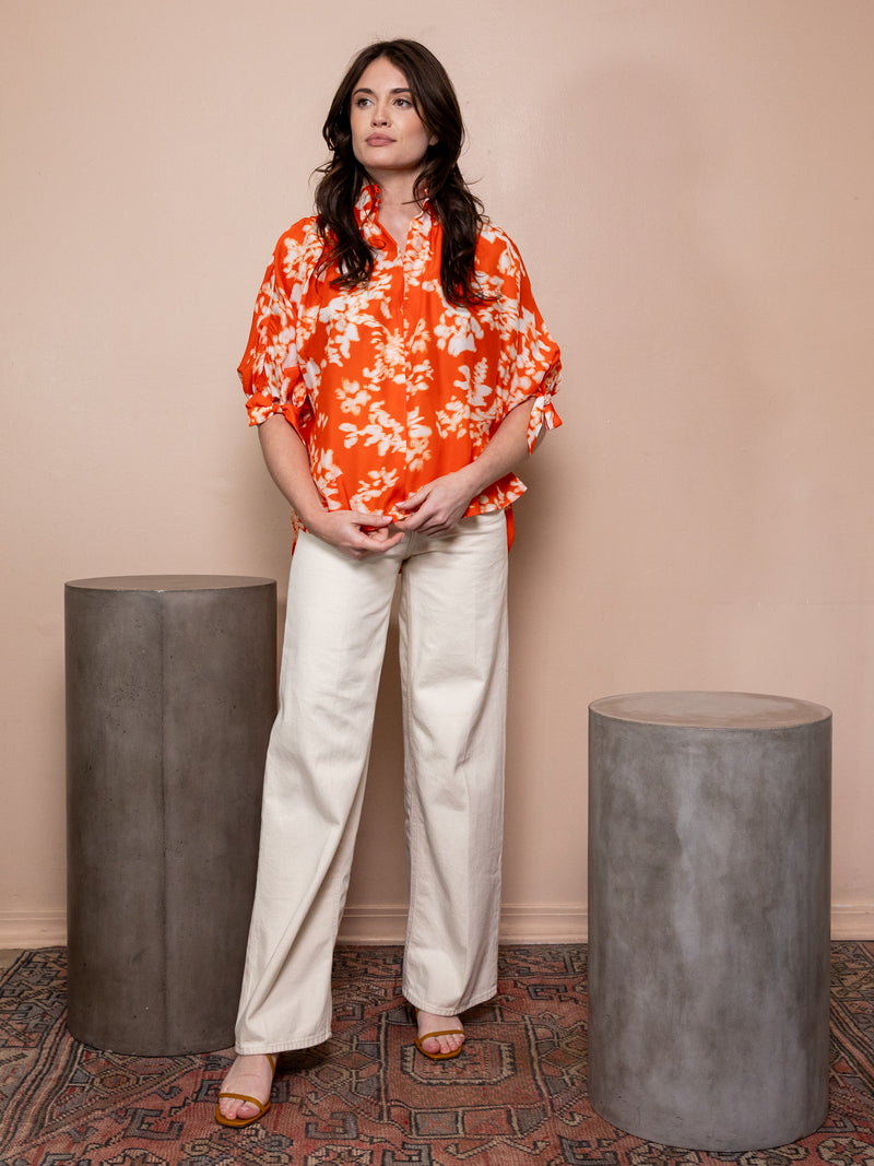 Woman wearing orange top with white pattern and white jeans against pink background.