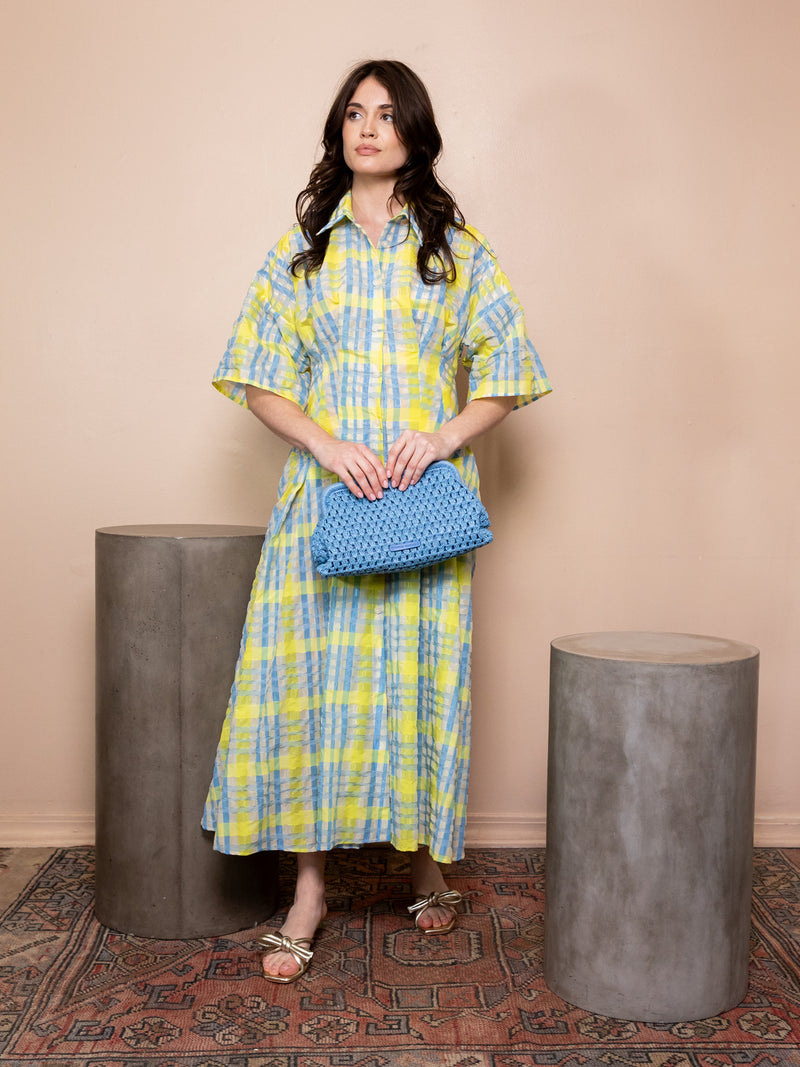 Woman wearing green and blue checkered dress and holding a blue bag against pink background.