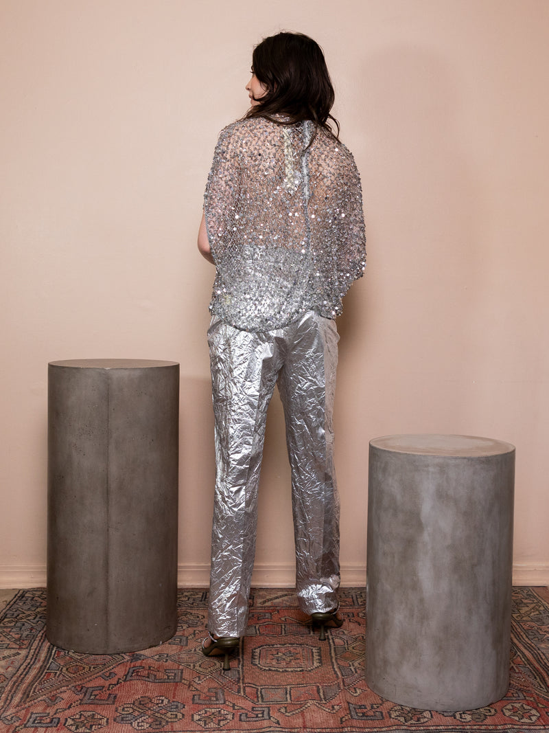 Woman wearing crinkled silver pants and silver sequin top against pink background