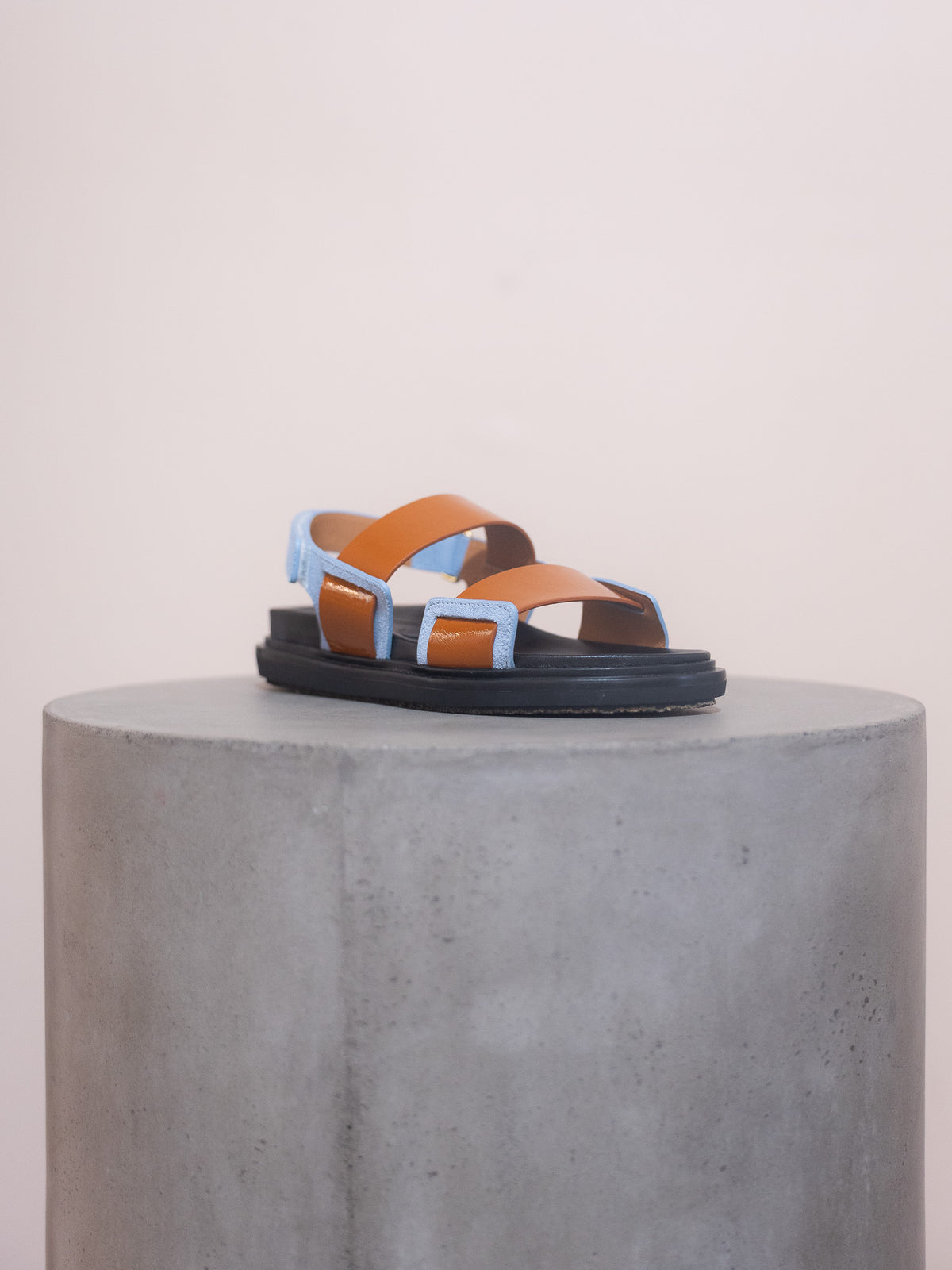 Brown and blue sandal on a gray pedestal against a light pink background