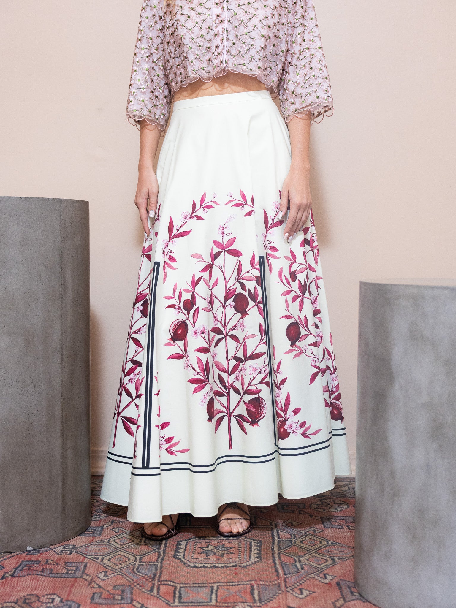 Person wearing a floral top and a long white skirt with pink floral patterns in a minimalistic setting.