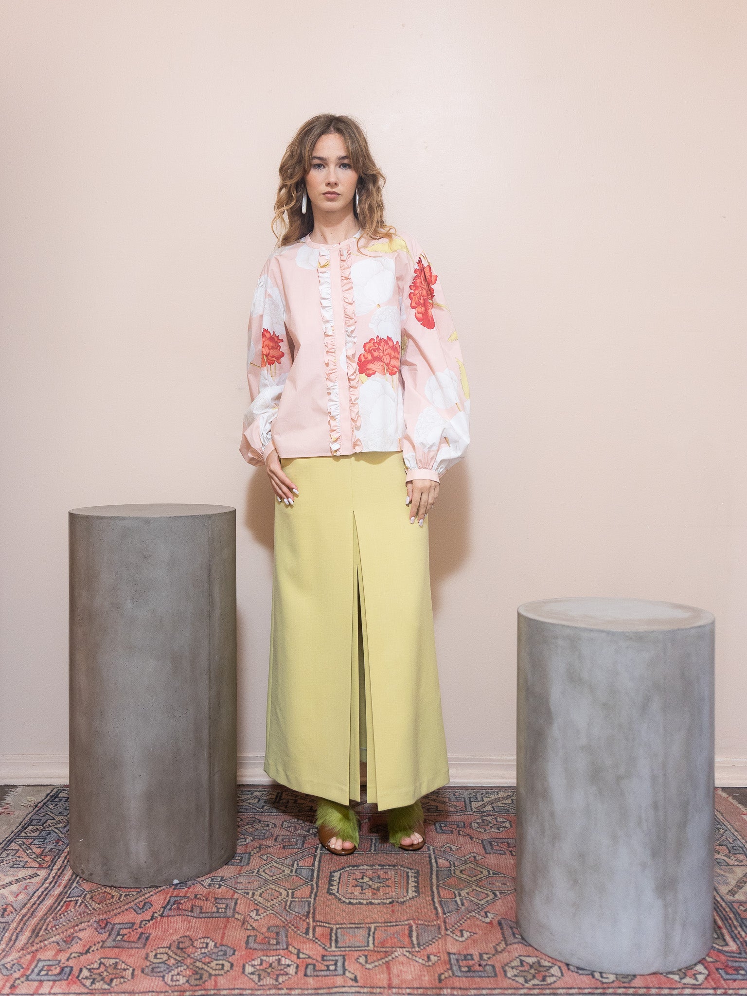 Woman in a floral blouse and green pants standing between two gray cylindrical objects on a patterned rug.