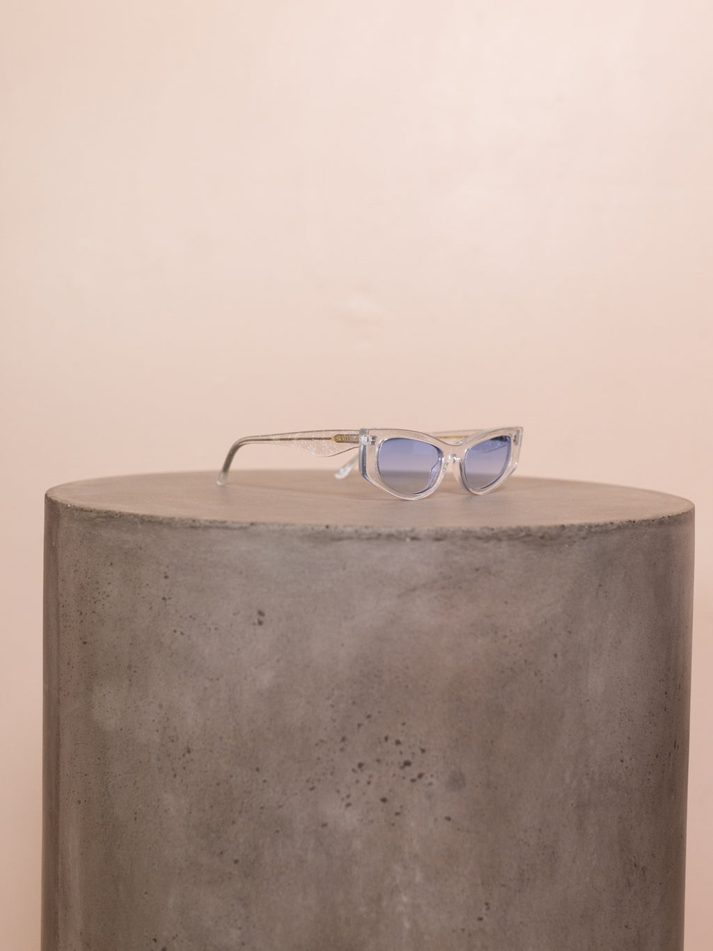 Clear sunglasses with blue lenses on pedestal against pink background.