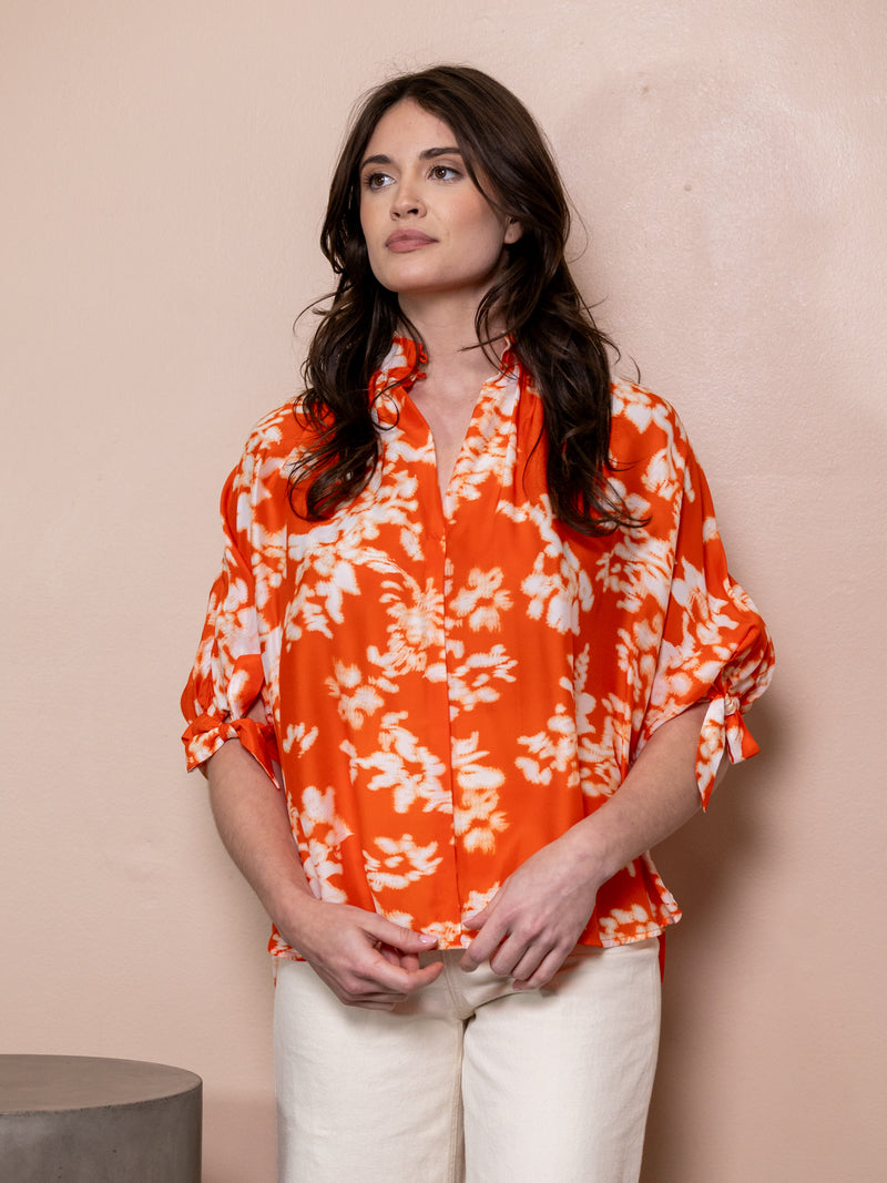 Woman wearing orange top with white pattern and white jeans against pink background.