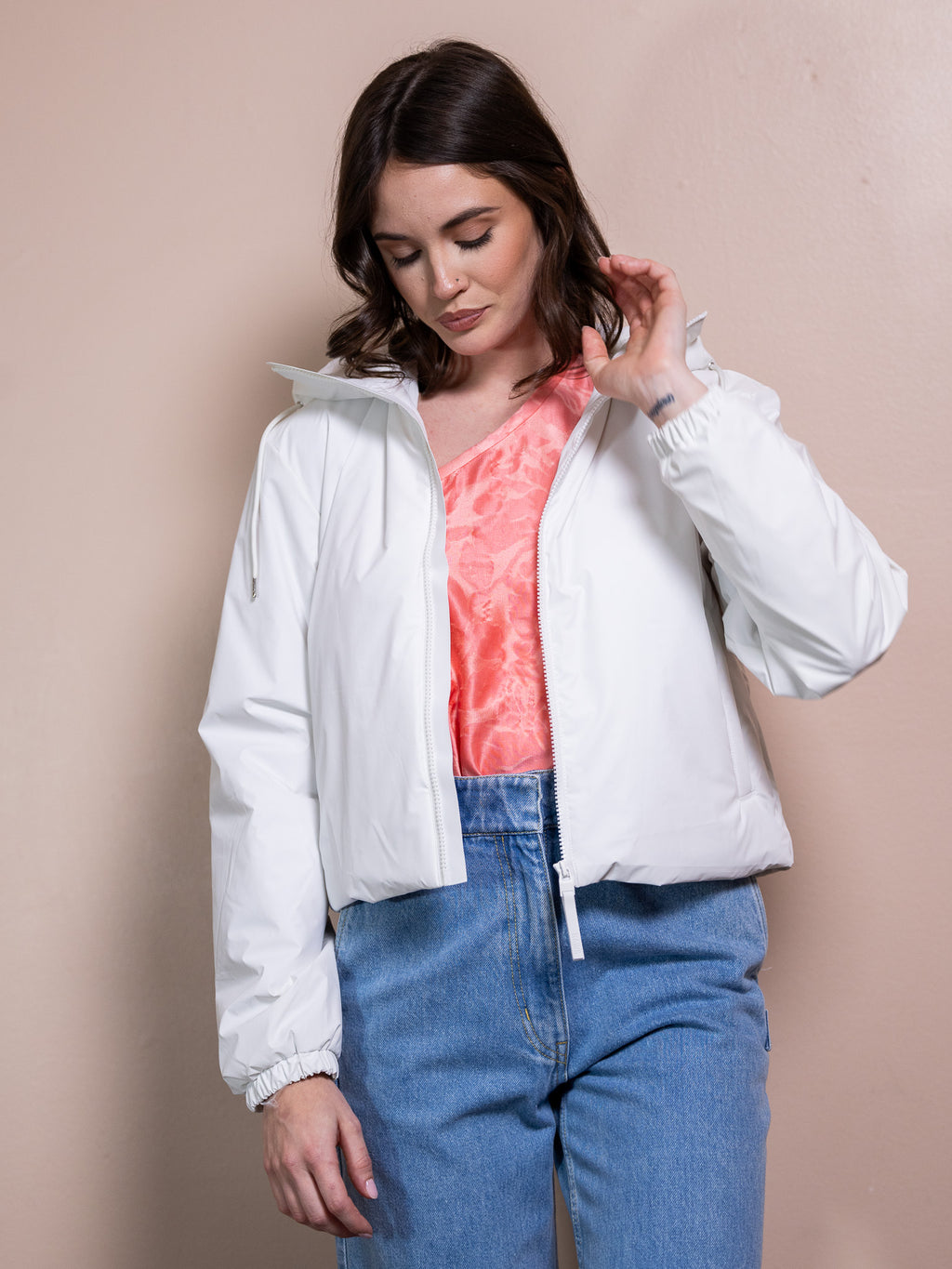 Woman in pink top, white rain jacket, and blue jeans against pink background