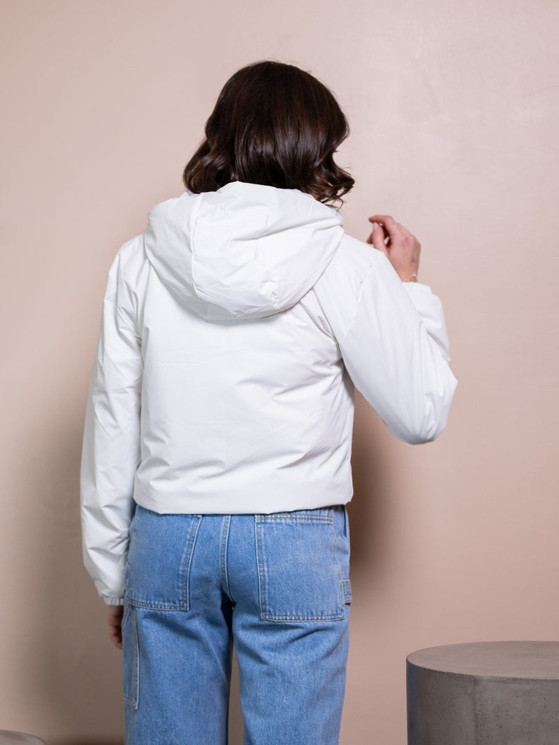 Woman in pink top, white rain jacket, and blue jeans against pink background