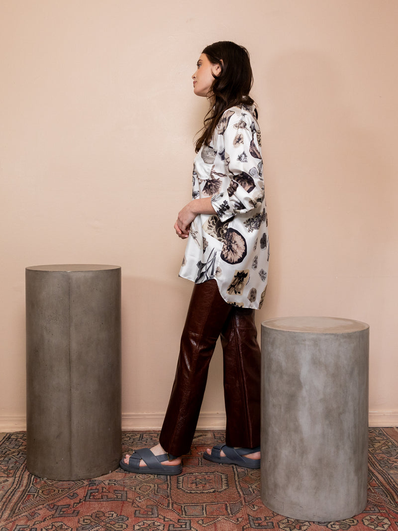 Woman wearing black and white floral collage top and brown leather pants against pink background.