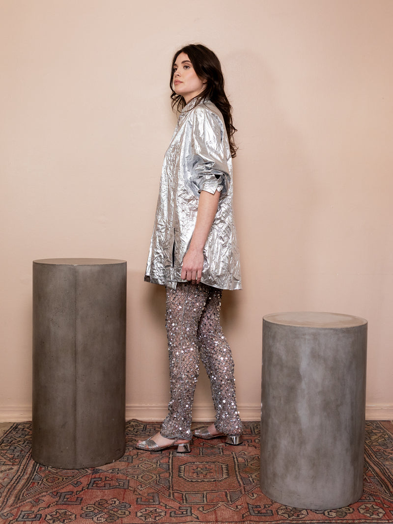 Woman wearing silver sequin pants and silver crinkled top against pink background.