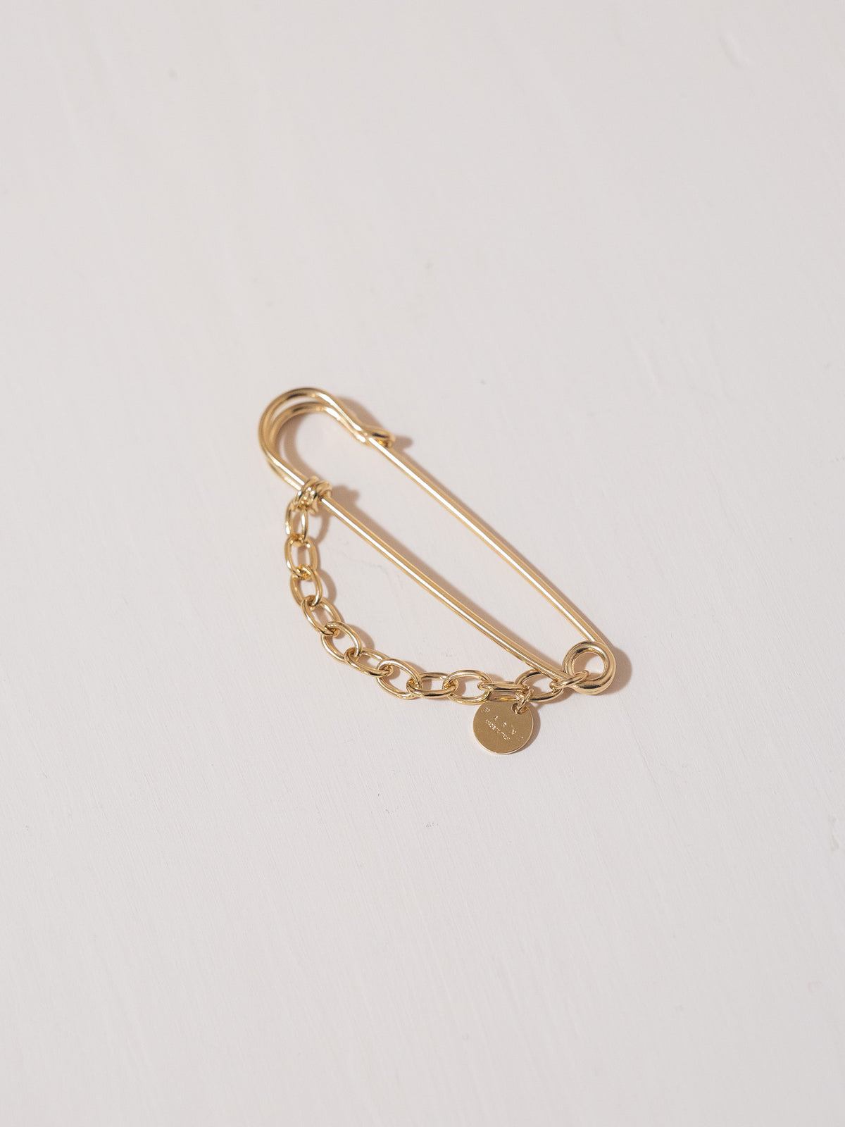 Gold safety pin with chain and round charm on a light gray background