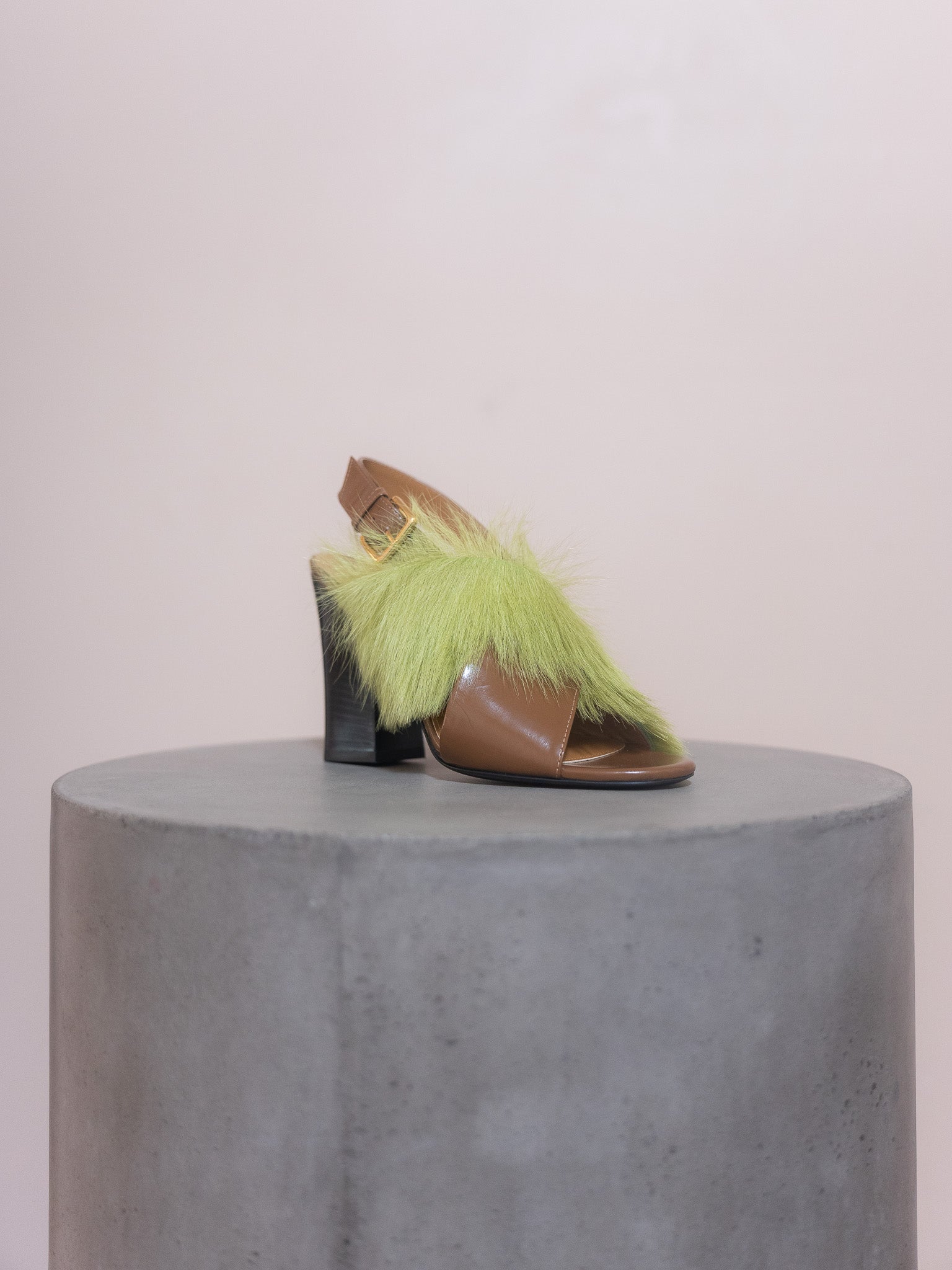 Brown open-toe shoe with green feathered strap on a gray pedestal against a light pink background