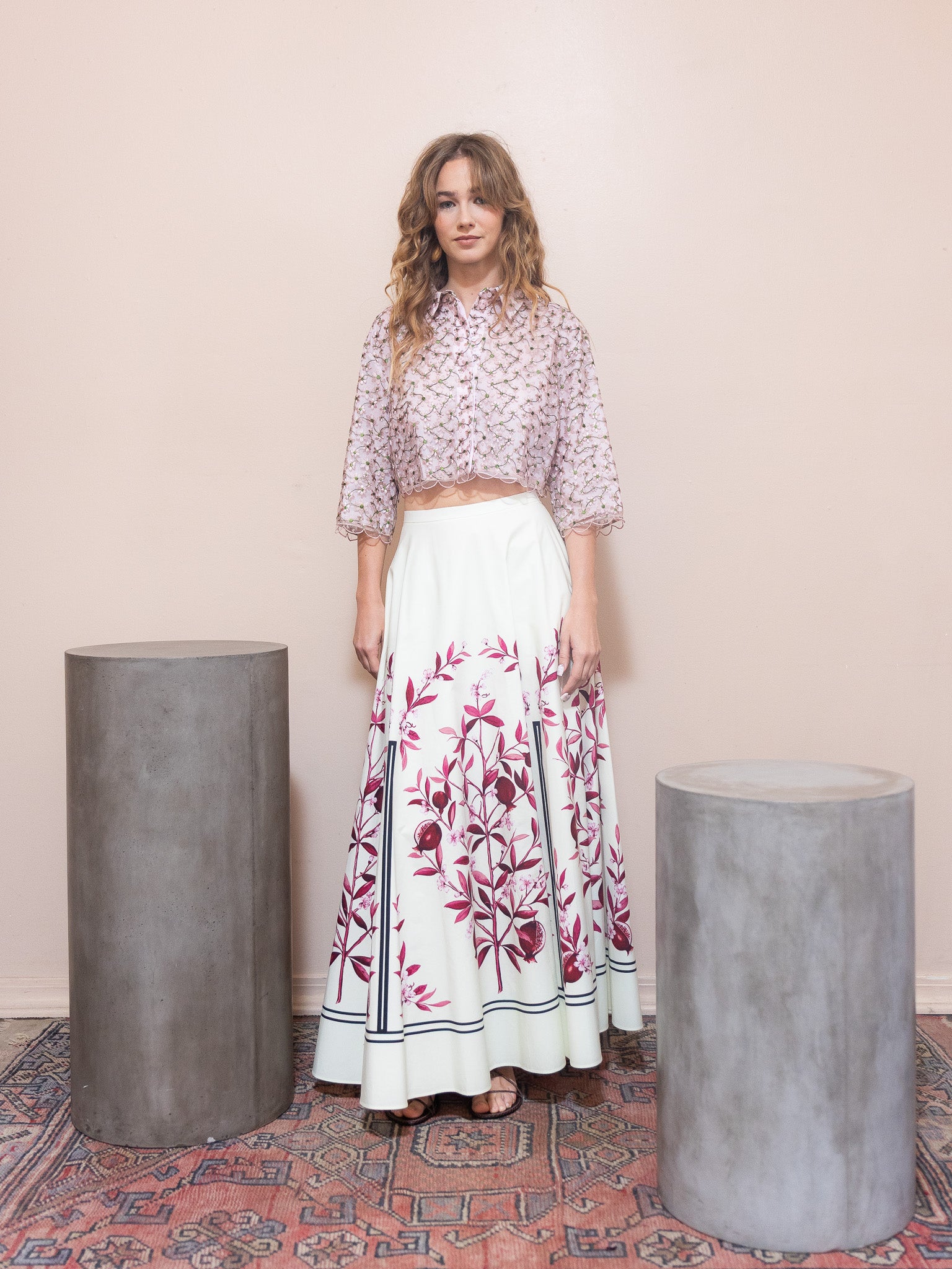 Woman wearing a floral top and white skirt with red patterns standing between two gray cylindrical objects on a patterned rug.