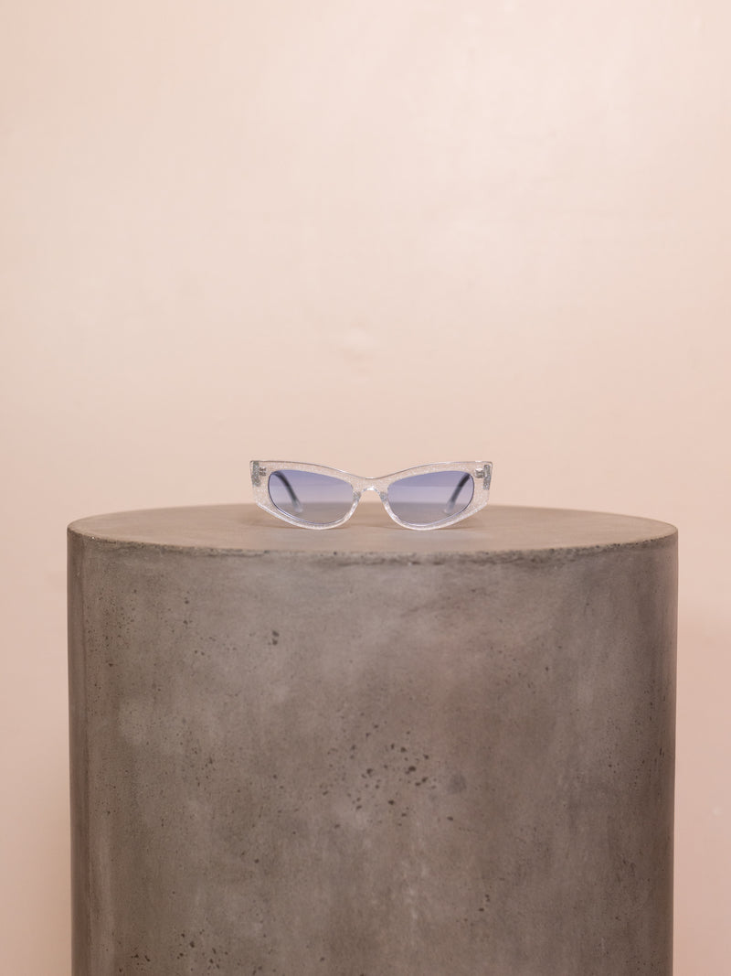 Clear sunglasses with blue lenses on pedestal against pink background.