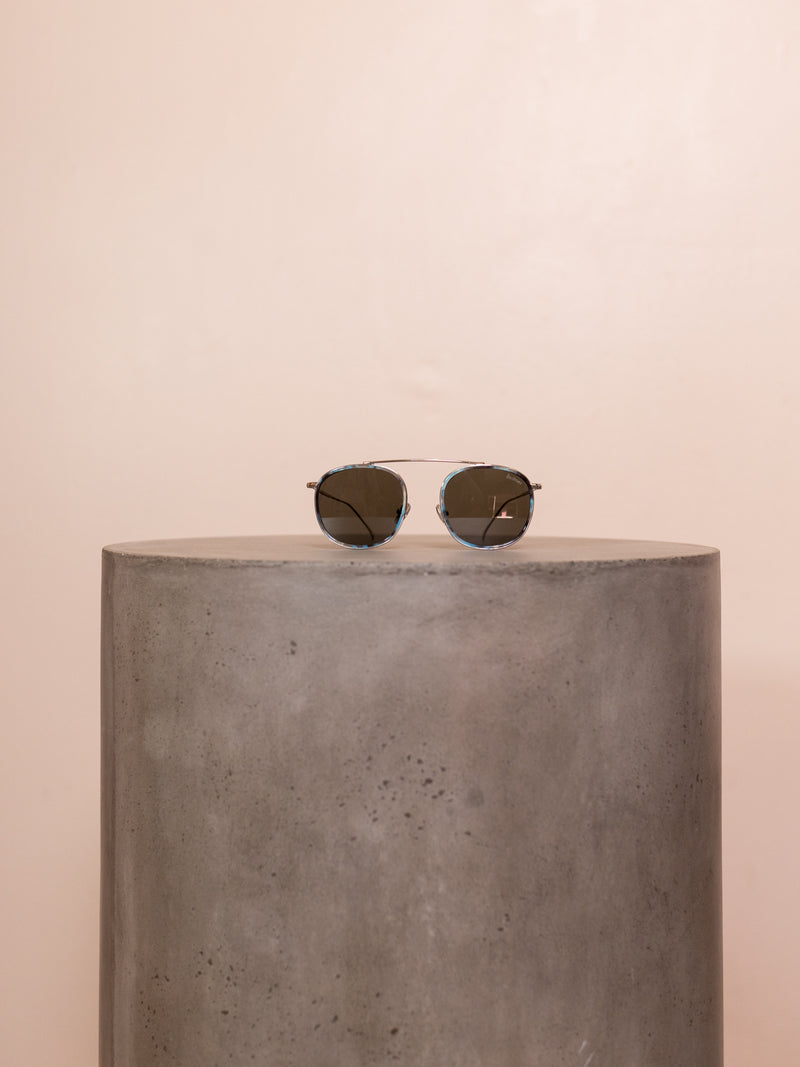 Wire framed sunglasses on pedestal against pink background.