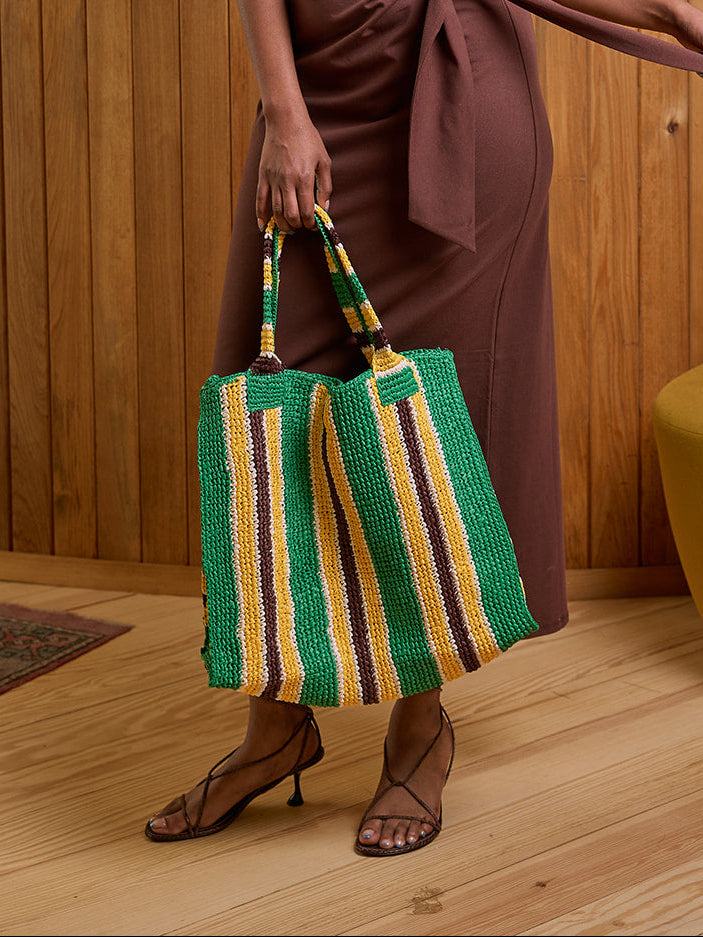 A woman stands in a wood-paneled room wearing a brown one-shoulder wrap dress, a cream headwrap, and brown strappy heels, while holding a green, yellow, and brown striped tote bag.