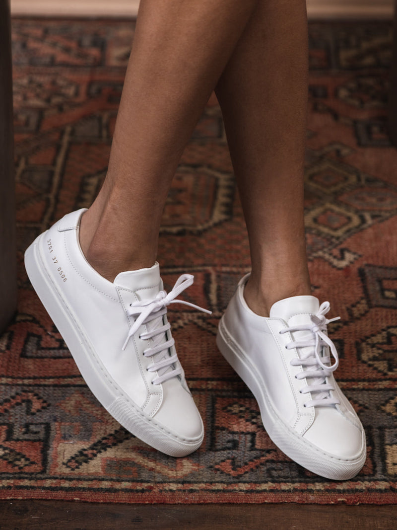 Woman wearing Common Projects white sneaker with white laces and gold embossed numbers on the side. Achilles Low Sneaker in White. Style Number: 3701