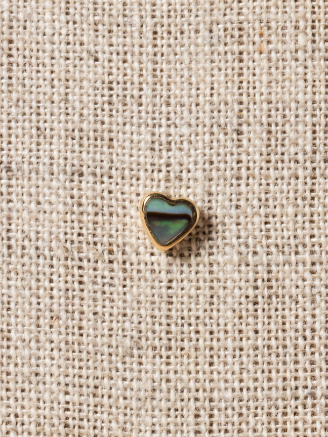 Heart-shaped stud earring with an abalone inlay, placed on a beige textured surface.