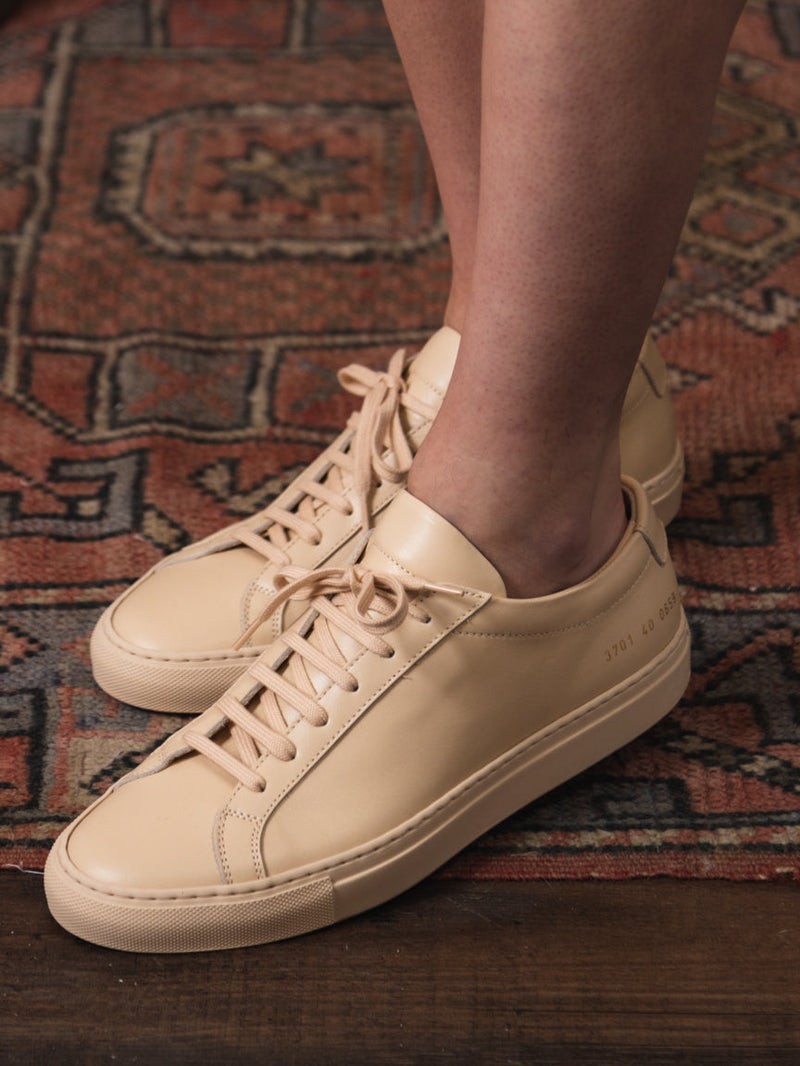 Woman wearing Common Projects tan sneaker with tan laces and gold embossed numbers on the side. Achilles Low Sneaker in Nude. Style Number: 3701